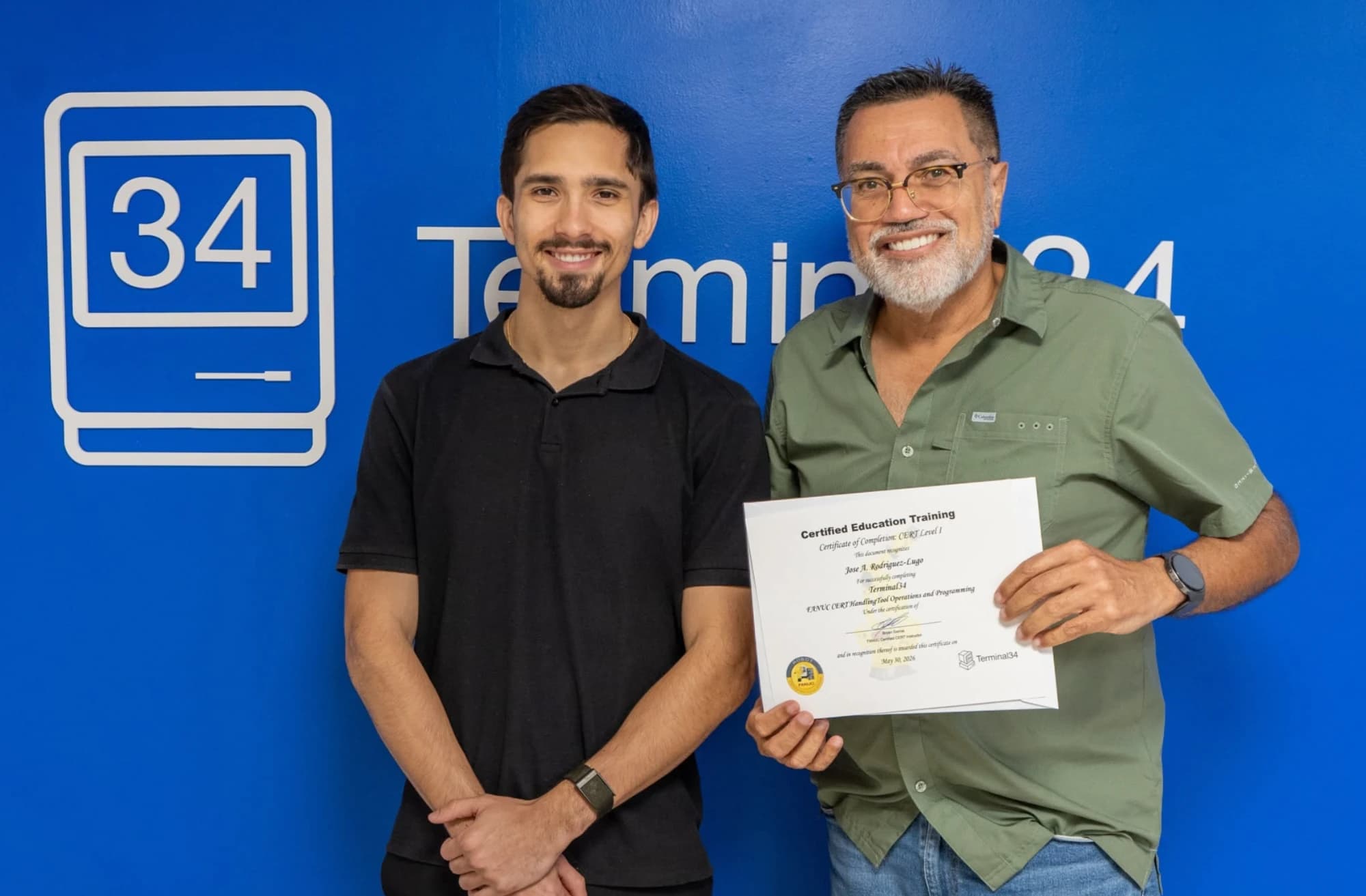 Graduado del curso de Robótica Industrial FANUC con camisa verde y su certificado junto al instructor