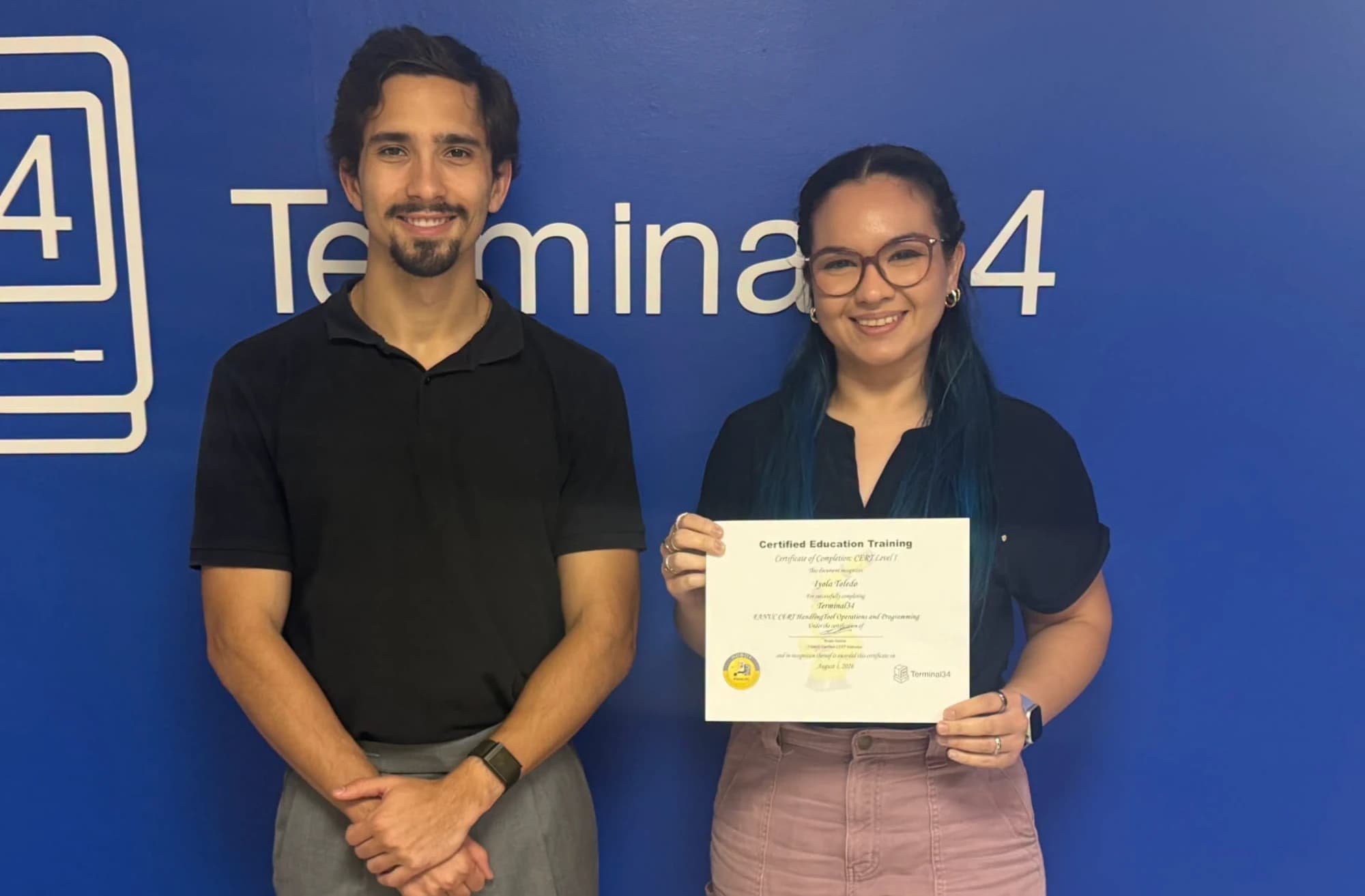 Graduada del curso de Robótica Industrial FANUC con su certificado junto al instructor, frente a la pared de Terminal34